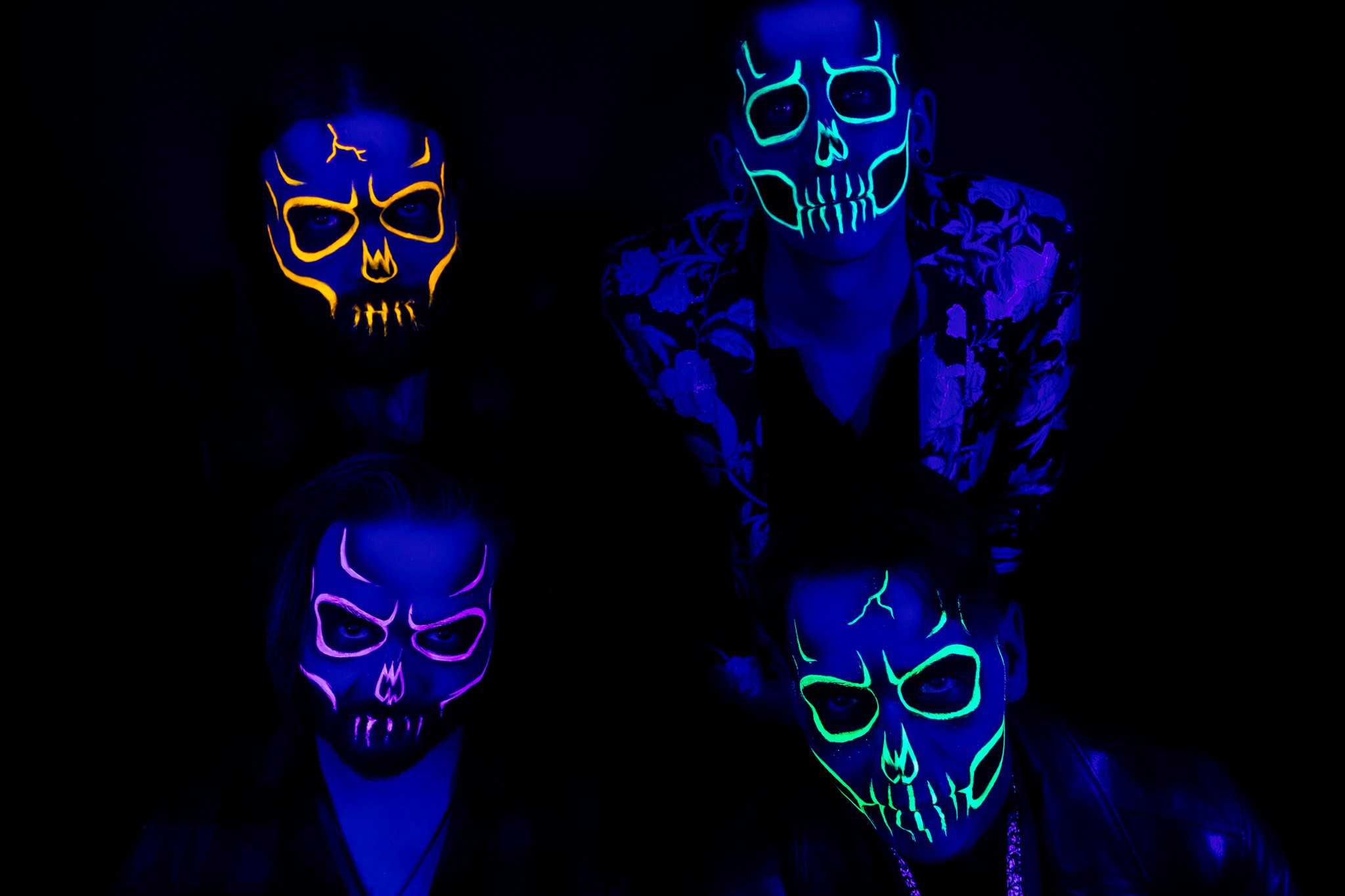 Against a black background, four men stand facing the camera, wearing UV-reactive skull facepaint