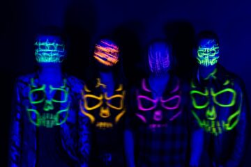 Against a black background, four men stand facing the camera, wearing UV-reactive skull facepaint, their faces blurred as they move, with superimposed images of their facepaint in front of them