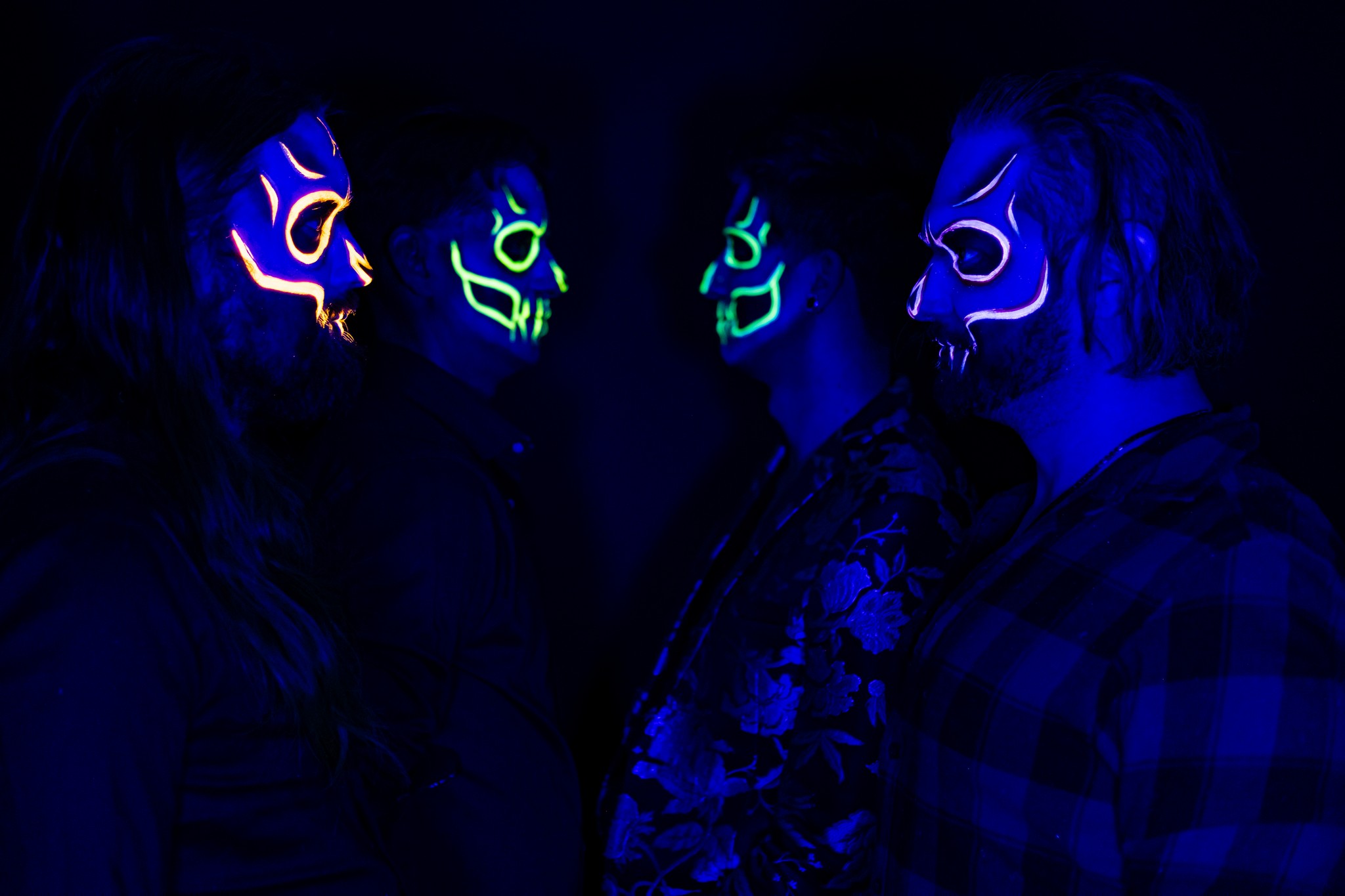 Against a black background, four men stand facing each other, two by two, wearing UV-reactive skull facepaint
